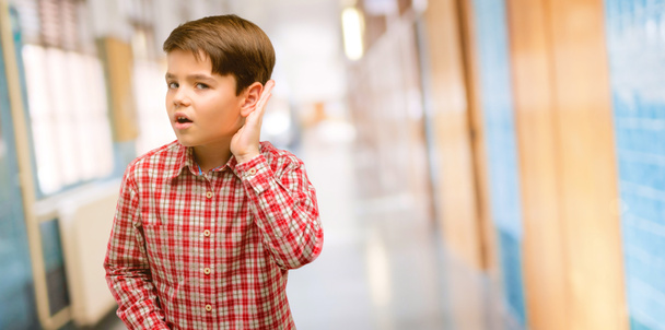 Красивый ребенок с зелеными глазами, держащийся за ухо, пытается слушать интересные новости, выражающие коммуникационную концепцию и сплетни в школьном коридоре.
 - Фото, изображение