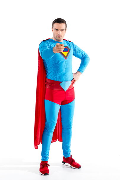 vista de longitud completa de superhéroe masculino seguro de pie con la mano en la cintura y apuntando a la cámara aislada en blanco
  - Foto, Imagen