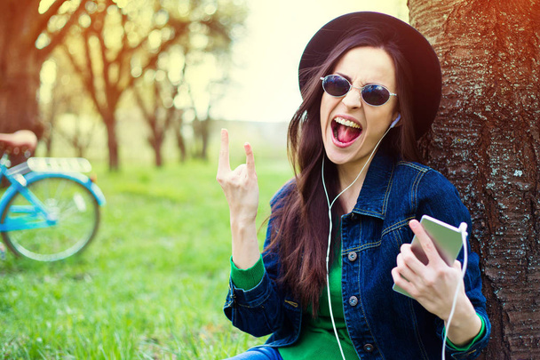 Casual woman in sunglasses and hat sitting on grass and listening music - Foto, imagen