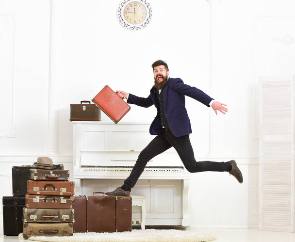 Macho attractive, elegant on cheerful face carries vintage suitcases, jumping. Man with beard and mustache in suit carries luggage, luxury white interior background. Long awaited vacation concept - Foto, Imagem
