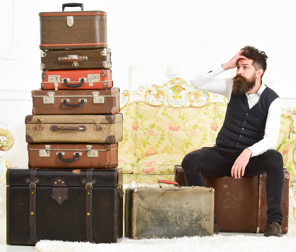 Man with beard and mustache packed luggage, white interior background. Macho elegant on tired face sits, exhausted at end of packing, near pile of vintage suitcases. Luggage and relocation concept - Foto, imagen