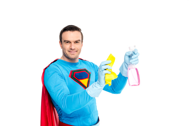knappe lachende superman bedrijf rag en spray fles met wasmiddel geïsoleerd op wit - Foto, afbeelding