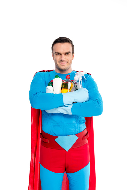 superman in rubber gloves holding cleaning items and smiling at camera isolated on white - Photo, Image