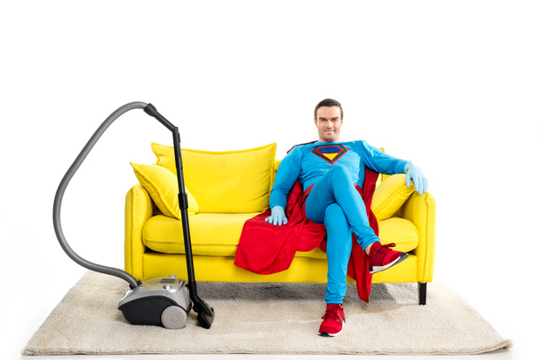 χαμογελώντας αρσενικό superhero στη λαστιχένια γάντια κάθεται στον καναπέ κοντά ηλεκτρική σκούπα και να βλέπουν τα φωτογραφικών μηχανών που απομονώνονται σε λευκό - Φωτογραφία, εικόνα
