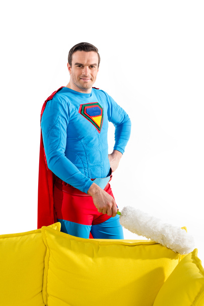 handsome male superhero cleaning couch with duster and smiling at camera isolated on white  - Photo, Image