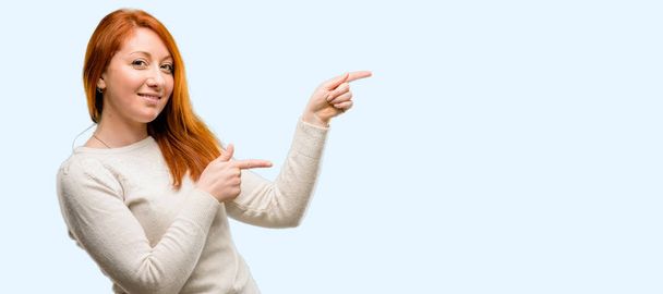 Beautiful young redhead woman pointing away side with finger isolated over blue background - Photo, Image