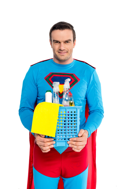 hombre guapo en traje de superhéroe sosteniendo suministros de limpieza y sonriendo a la cámara aislada en blanco
 - Foto, imagen