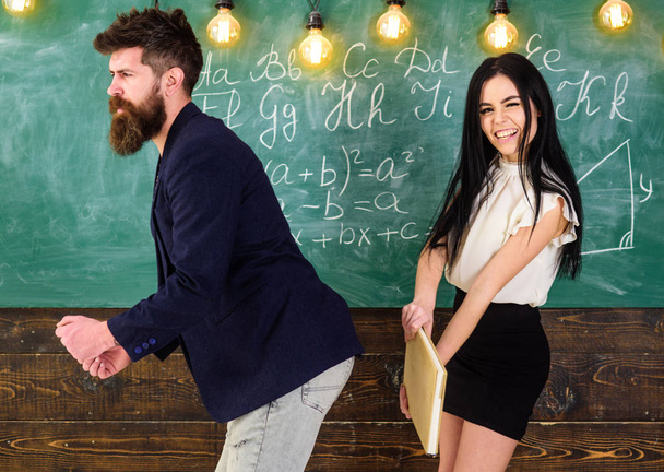 Student lady slapping on teachers buttocks with book. Girl on smiling face having fun while punishes teacher. Man with beard punished by sexy student, chalkboard on background. Role games concept - Фото, изображение