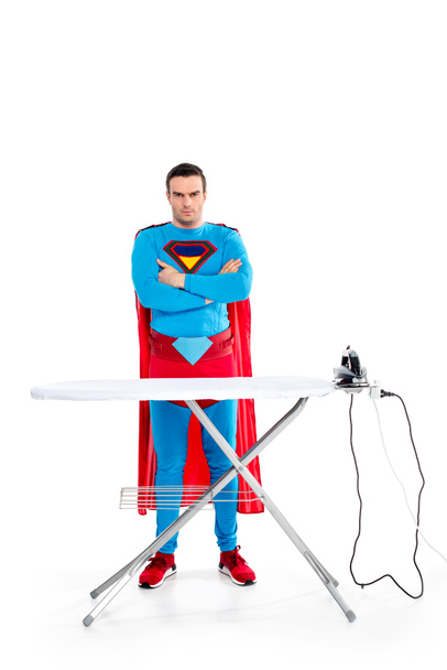 handsome superman standing with crossed arms near ironing board and looking at camera on white - Foto, Bild