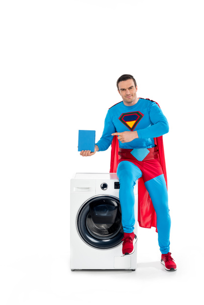 smiling male superhero sitting on washing machine and pointing with finger at soap powder on white - Photo, Image