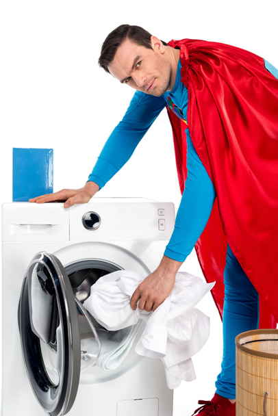 superman washing clothes in washing machine and looking at camera on white  - Photo, Image