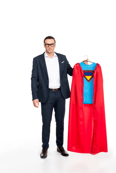 businessman in eyeglasses holding hanger with superhero costume and looking at camera isolated on white  - Photo, Image