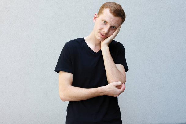 fashion, style, emotions and people concept - Close up portrait of a redhead of a beautiful manly guy with freckles on a gray background black T-shirt. holding head with hand being tired - 写真・画像