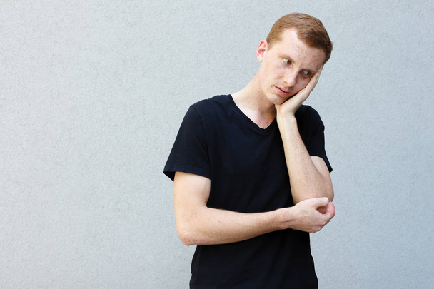 fashion, style, emotions and people concept - Close up portrait of a redhead of a beautiful manly guy with freckles on a gray background black T-shirt. holding head with hand being tired - Φωτογραφία, εικόνα