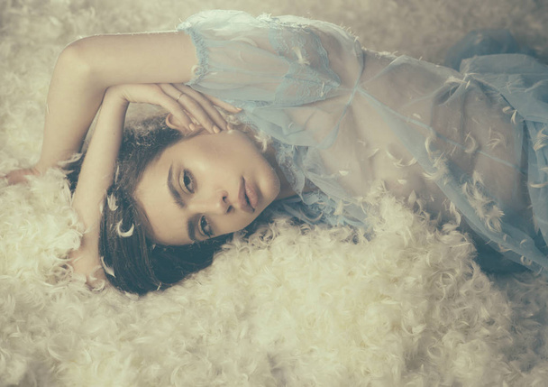 Beautiful girl in blue nightgown falling asleep. Young female with brunette hair lying on soft white feathers, tenderness and airiness concept - 写真・画像