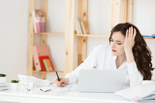 Устала красивая предпринимательница, держащаяся за голову во время работы с компьютером и некоторыми деловыми документами в ярком офисе
 - Фото, изображение