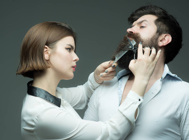 Barbershop or hairdresser concept. Woman hairdresser cuts beard with scissors. Guy with modern hairstyle visiting hairdresser. Man with long beard, mustache and stylish hair, grey background - Foto, Imagem