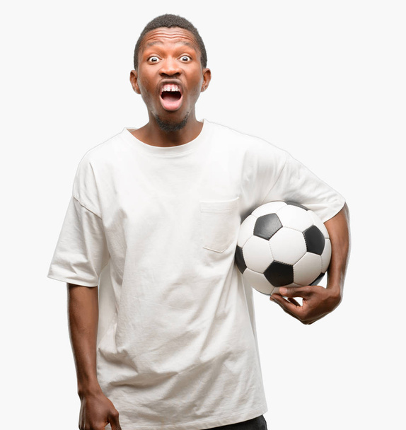 Afryki czarny człowiek trzymanie piłki nożnej, stresujące, przerażony w panice, krzycząc zdenerwowany i sfrustrowany. Nieprzyjemne gest. Irytujące pracy doprowadza mnie do szału - Zdjęcie, obraz