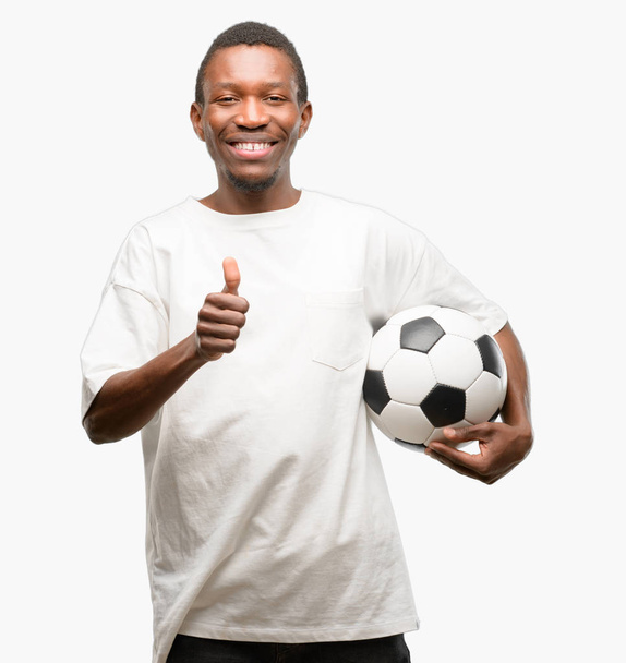 広く示す笑みを浮かべてサッカー ボールを保持しているアフリカの黒人男性の親指カメラのような式および承認するジェスチャを - 写真・画像