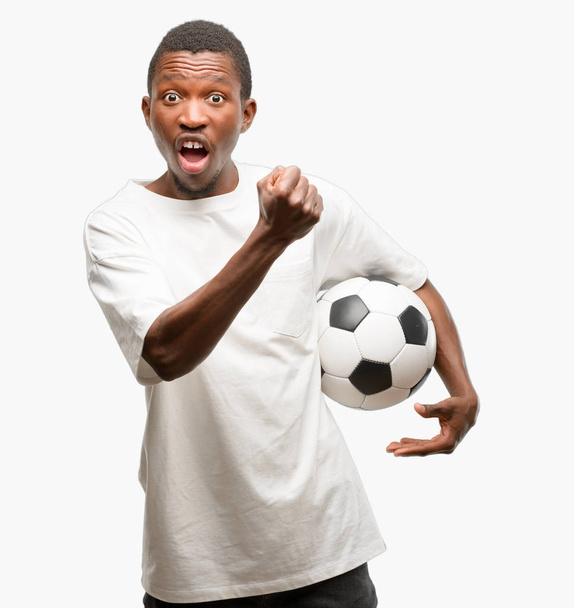 Αφρικανική μαύρο άνθρωπος που κρατά μπάλα ποδοσφαίρου, ενοχλημένος και θυμωμένος εκφράζοντας αρνητικά συναισθήματα, ενοχλημένος με κάποιον - Φωτογραφία, εικόνα