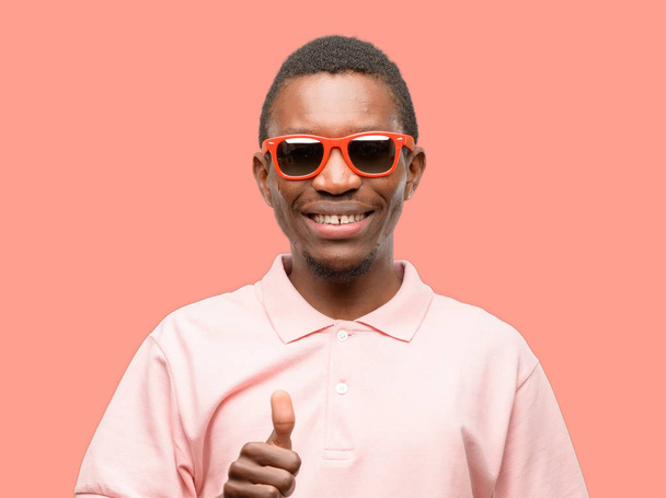 Afrikaanse zwarte man dragen van een zonnebril glimlachend in grote lijnen weergegeven: duimschroef opwaarts gebaar camera, expressie van en goedkeuring - Foto, afbeelding
