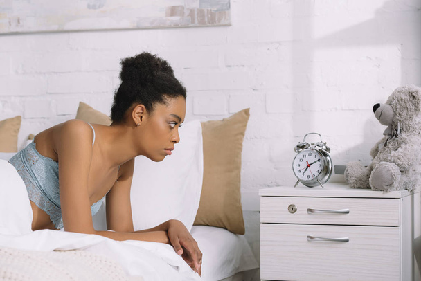 upset african american woman looking at alarm clock while lying in bed - Foto, Imagem