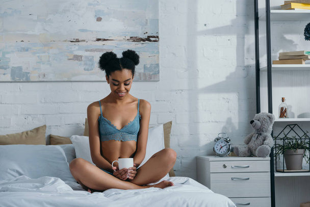 smiling african american girl in underwear with cup of coffee sitting on bed in bedroom in morning  - Photo, Image