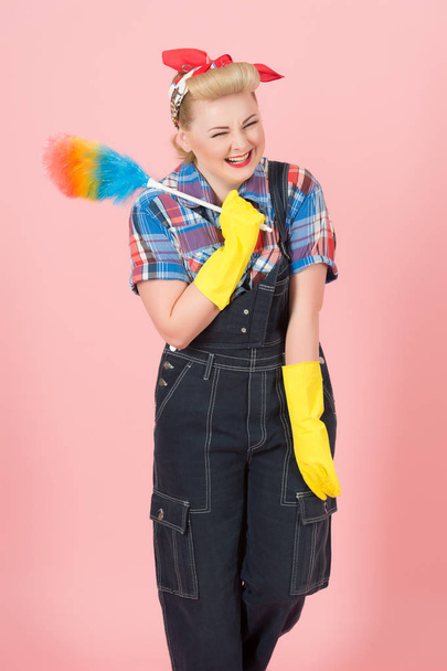 Housekeeper blonde girl in denim with colored duster and happy closed eyes. Blonde girl in american style with curls and red head scurf very smiled while cleaning. - Фото, изображение