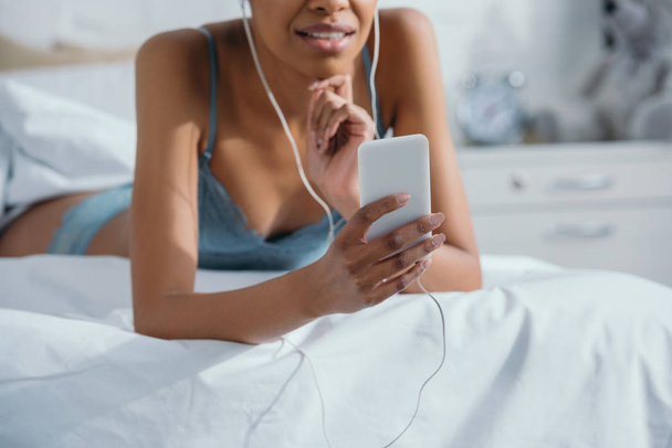 cropped view of african american girl listening music with earphones and using smartphone on bed - Zdjęcie, obraz