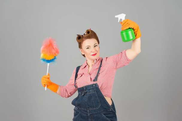 Girl in pin up styled with soft colorful duster and green spray bottle on grey background. Denim girl in orange gloves with brunette curls. Brunette woman ready for cleaning duster and spray bottle - Zdjęcie, obraz