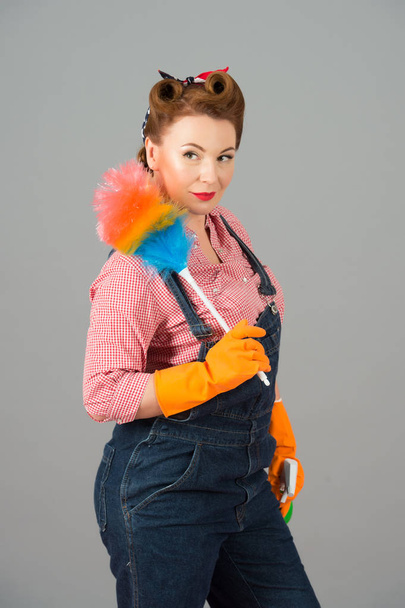 Cleaning lady with colored duster and orange rubber gloves in pin-up style. Brunette curl retro styled housekeeper on grey background - Fotoğraf, Görsel