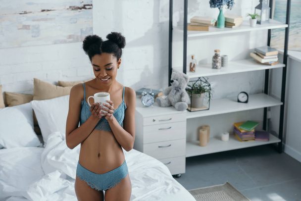 beautiful smiling african american woman in underwear standing with cup of coffee in bedroom in the morning   - Φωτογραφία, εικόνα