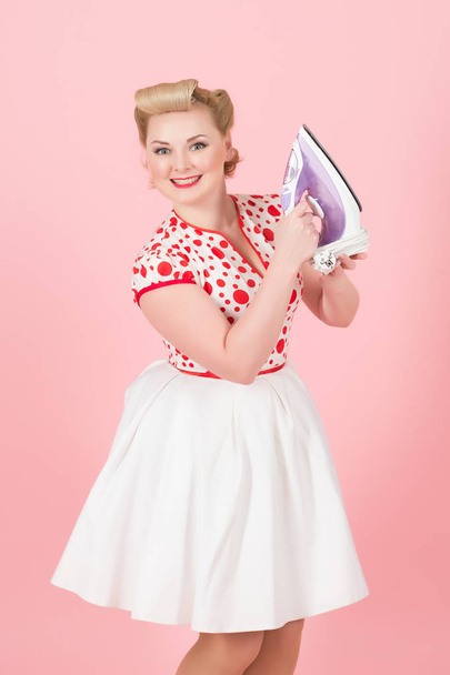 Young pin-up styled girl holds iron on pink background. Beautiful pin-up blonde woman with curls prepare iron for work isolated in studio. Blonde lady happy to have iron in hands. Pretty blonde girl - Fotografie, Obrázek