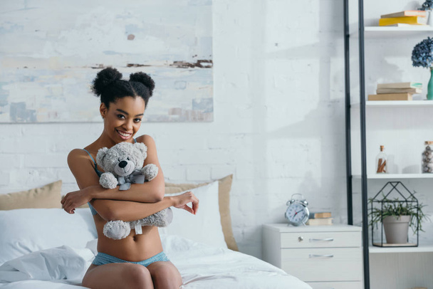 smiling african american girl hugging teddy bear while sitting on bed in morning  - Fotoğraf, Görsel