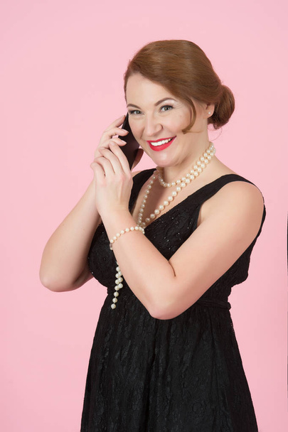 Beautiful brunette woman speak over smart phone. Cheerful woman holds telephone. Woman in black with perls on pink background. Lady in black dress happy talking over the smart phone. - Photo, image