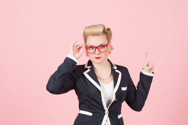 blonde girl in jacket and red glasses pointing up on pink background. woman with blonde curves holds the glasses on face and warning up by finger on place for texting. serious teacher asks attention - Photo, Image