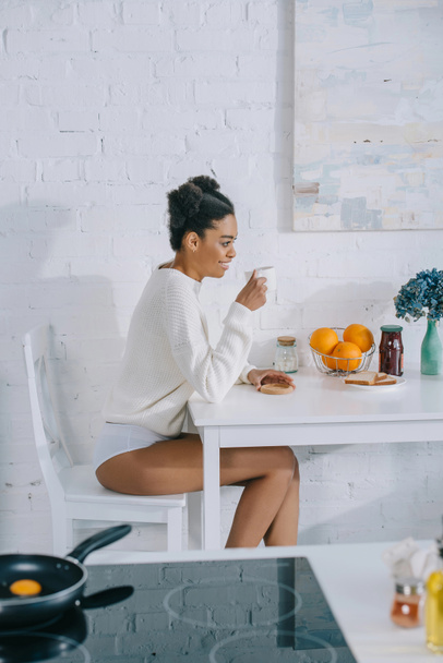 вид сбоку улыбающейся молодой женщины с чашкой утреннего кофе дома
 - Фото, изображение