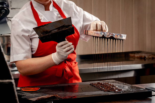 eine Konditorin mit roter Uniform und weißen sterilen Handschuhen macht ein Set bunter Pralinen aus Vollmilchschokolade auf einem Tisch. - Foto, Bild