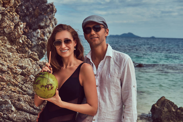 attraktives Paar, hält Kokosnuss mit Schlauch, steht an einem Strand in der Nähe großer Riffsteine, genießt einen Urlaub auf einer wunderschönen Insel.  - Foto, Bild
