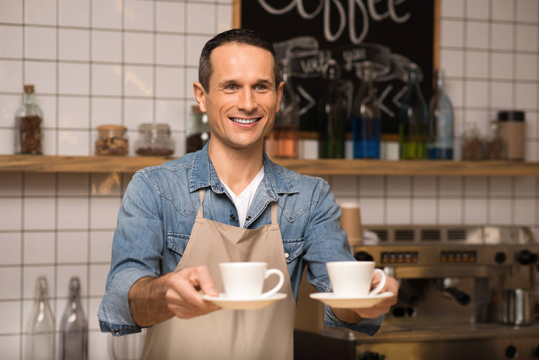 portrait of smiling barista holding two cups of coffee and looking away - Fotó, kép