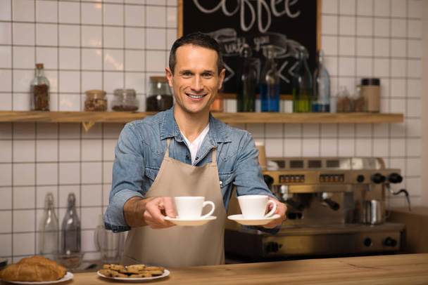 portrait of smiling young waiter holding two cups of coffee and looking at camera - Foto, Bild