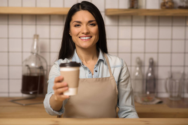 portrait of smiling waitress holding coffee to go in paper cup and looking at the camera - Фото, зображення