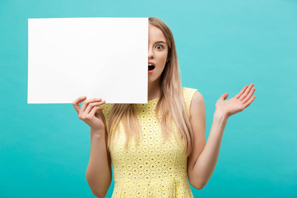 Retrato de una joven rubia asombrada sosteniendo un letrero en blanco con espacio de copia sobre fondo azul del estudio. Mostrando la cara sorprendida sorpresa
. - Foto, Imagen