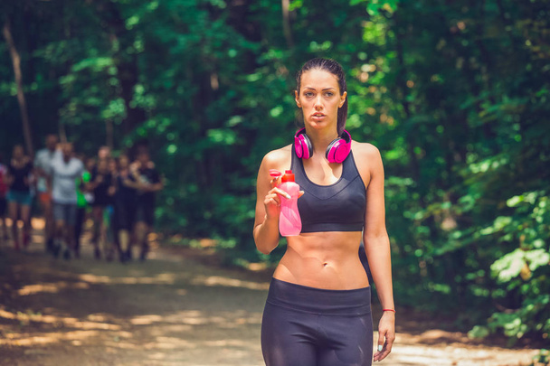 Young athlete walking in woods and holding bottle of water. Group of runners in the background. - Zdjęcie, obraz