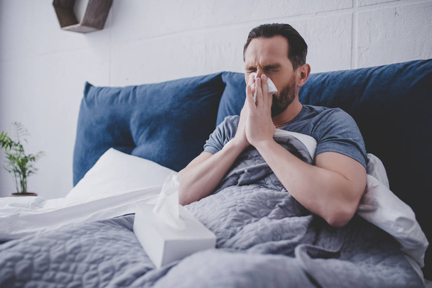 Mid adult man sneezing in napkin sitting on the bed at home - Foto, Imagen