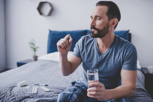 caucasian man holding pill and glass of water sitting on the bed - Foto, immagini