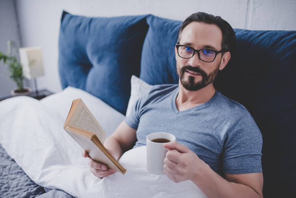 Man on glasses reading book and holding cup of tea in bed, looking at the camera - Foto, Imagen