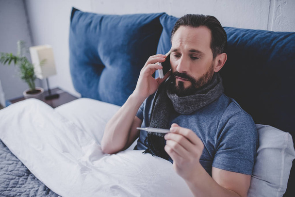 portrait of sick man holding thermometer and calling a doctor while sitting on the bed - Foto, imagen