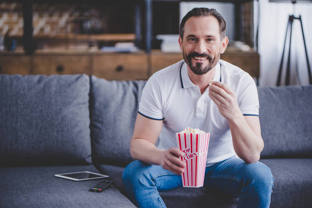 smiling man sitting on the sofa, watching tv and eating popcorn at home - Valokuva, kuva