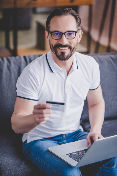 bearded man shopping online with credit card and using laptop at home while looking at the camera - Fotoğraf, Görsel